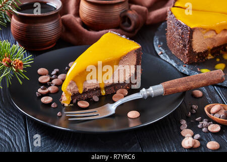 Close-up di un pezzo di cioccolato mango cheesecake servita su una piastra nera con forcella di dessert e scaglie di cioccolato. abete e tazze di caffè su un Foto Stock