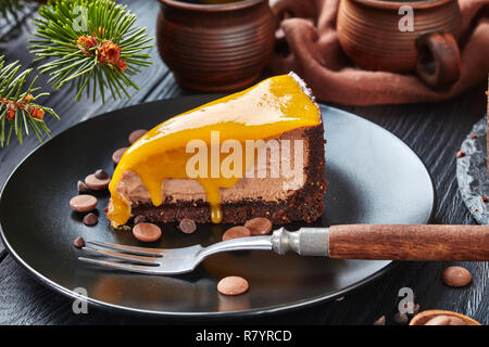 Close-up di un pezzo di cioccolato mango cheesecake servita su una piastra nera con forcella di dessert e scaglie di cioccolato. abete e tazze di caffè a th Foto Stock