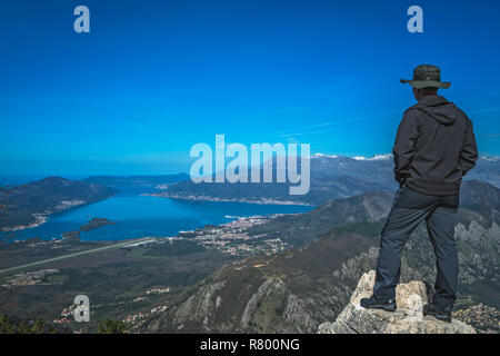 Turistico con un cappello in piedi su un grande masso e ammirando il panorama mozzafiato della Baia di Kotor in Montenegro come visto dalla strada di Lovcen N Foto Stock