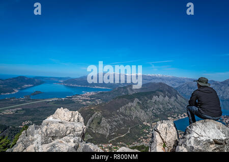 Turistico con un cappello seduto su un grosso masso e ammirando il panorama mozzafiato della Baia di Kotor in Montenegro come visto dalla strada di Lovcen Na Foto Stock