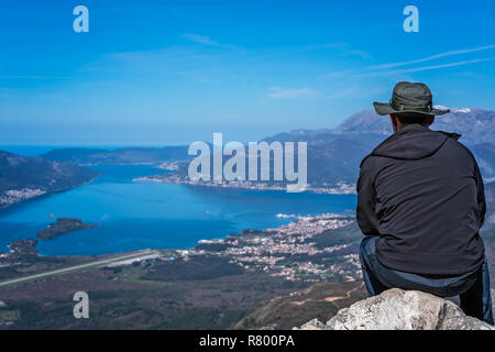 Turistico con un cappello seduto su un grosso masso e ammirando il panorama mozzafiato della Baia di Kotor in Montenegro come visto dalla strada di Lovcen Na Foto Stock