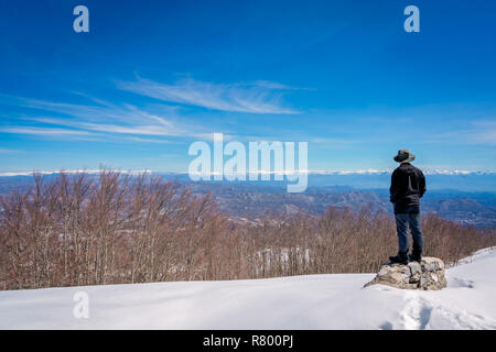 Voce maschile turistica prendendo indossando un cappello in piedi su una roccia e ammirando la splendida montagna paesaggio invernale panorama visibile dalla cima del monte Lovcen Foto Stock