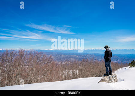 Voce maschile turistica prendendo indossando un cappello in piedi su una roccia e ammirando la splendida montagna paesaggio invernale panorama visibile dalla cima del monte Lovcen Foto Stock