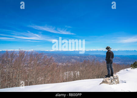 Voce maschile turistica prendendo indossando un cappello in piedi su una roccia e ammirando la splendida montagna paesaggio invernale panorama visibile dalla cima del monte Lovcen Foto Stock