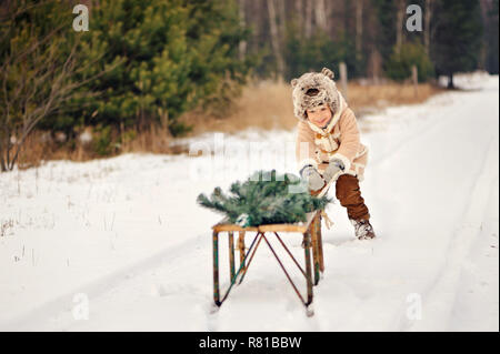 Un bambino decorare un albero di Natale all'aperto. Il ragazzo si blocca red bolle e mangia gingerbreads. Egli è vestito con un cappotto di pelle di pecora, nel verde dello sfondo Foto Stock