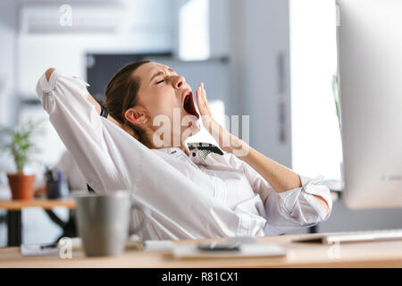 Assonnato giovane donna vestita in camicia seduto al suo posto di lavoro presso l'ufficio, sbadigli Foto Stock