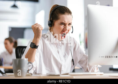 Arrabbiato giovane donna indossa cuffie con microfono vestito in camicia seduto al suo posto di lavoro presso l'ufficio, urlando Foto Stock