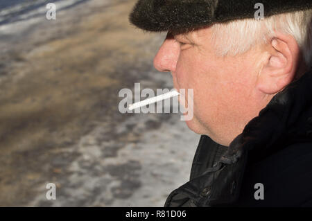 Un anziano grigio-dai capelli uomo in abbigliamento invernale tenendo una sigaretta in bocca Foto Stock