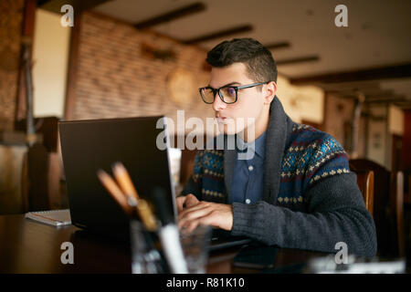 Bello il freelance proprietario in bicchieri e inverno maglione diligentemente lavorando sul computer portatile in cafe. L'uomo la digitazione sulla tastiera e cerca un nuovo lavoro su internet al coffee shop. Il concetto di business Foto Stock