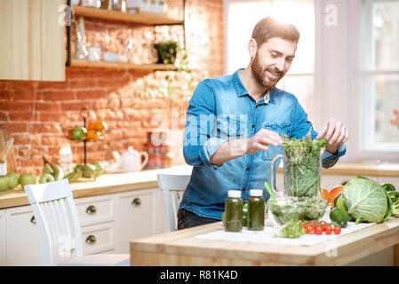 Uomo bello il riempimento di Blender con verde cibo crudo, rendendo frullato vegetariano sulla cucina a casa Foto Stock