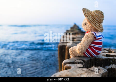 Orsacchiotto di peluche in vacanza estiva sedersi sulla fila di pennelli in legno, usura cappello di paglia e striato sailor suit, prendere uno sguardo verso il blu acqua orizzonte (copia spazio) Foto Stock