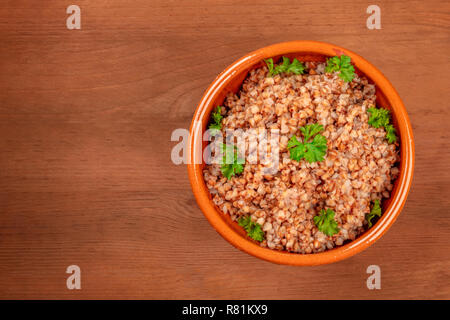 Cuocere il grano saraceno in un recipiente di terracotta, guarnite con prezzemolo fresco in foglie, al buio su un rustico tavolo in legno con spazio di copia Foto Stock