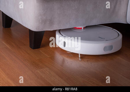 Robot bianco aspirapolvere gira in un angolo sotto il divano su legno parquet. Smart moderna tecnologia di pulizia le pulizie. Foto Stock