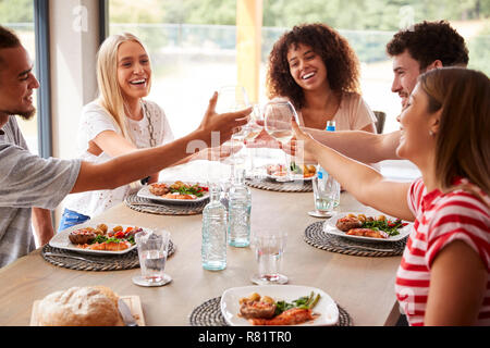 Multi etnico gruppo di cinque giovani felici amici adulti ridendo e sollevamento di occhiali per toast durante una cena Foto Stock