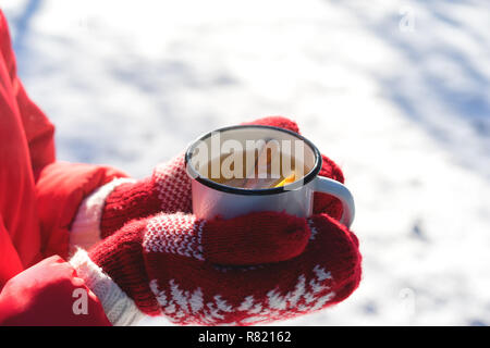 Mani in maglia Mittens holding fumante tazza di tè caldo su nevoso inverno mattina all'esterno. La donna trattiene accogliente rosso festoso la tazza con una bevanda calda mattina di Natale Foto Stock