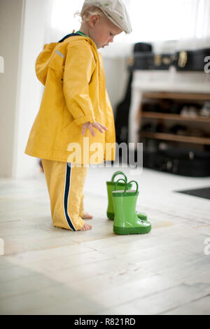 Giovani toddler in un impermeabile guardando verso il basso in corrispondenza di una coppia di verde stivali di gomma all'interno di una casa. Foto Stock