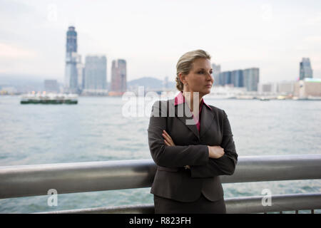 Mid-adulto imprenditrice sta in piedi in una ringhiera che si affaccia su una città portuale con le braccia incrociate. Foto Stock