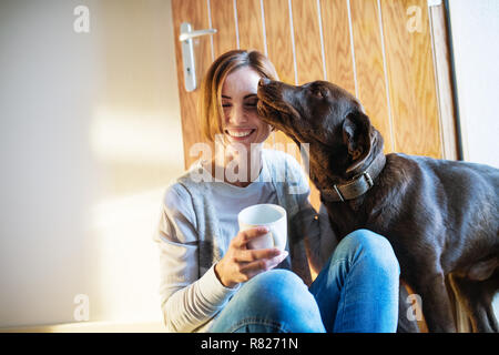 Una giovane donna seduta al chiuso sul pavimento a casa, giocando con un cane. Foto Stock