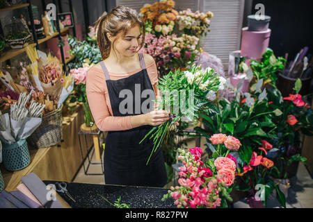 Allegro giovane donna stand e combinare i fiori. Ella fa bouquet. Fioraio sorriso. Stand ella nella stanza piena di fiori e piante. Foto Stock