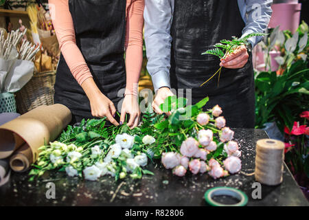Vista di taglio del giovane uomo e donna in nero grembiule stare a tavola. Essi fanno mazzi da bianco e luce rosa rose. Egli addd alcune felci frondoso. Foto Stock