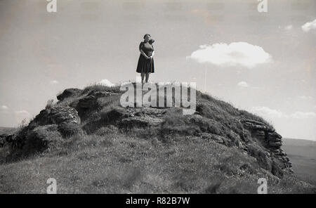 1950, storico, una signora walker in piedi al di fuori su una coperta di erba picco roccioso o Tor di Dartmoor Devon, Inghilterra, Regno Unito. Uno degli aspetti più caratteristici del moro, questi picchi rocciosi sono disponibili in molte forme, anche se la maggior parte sono composte di granito. Foto Stock