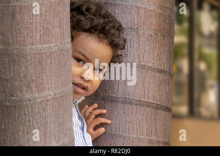 Un curioso ragazzino picchi presso la telecamera mentre tra i due alberi. Fuori sul marciapiede, un ragazzino curioso si nasconde dietro alberi mentre egli guarda. Foto Stock