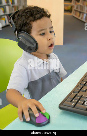 Un Littell ragazzo seduto davanti a un computer tenendo un mouse e cuffie sembra sorpreso da ciò che vede. Foto Stock