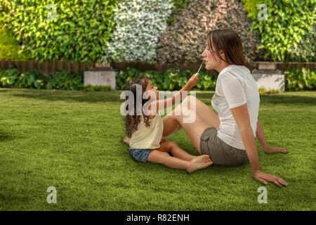 Madre e figlia di specchio tempo di gioco. Tre-anno-vecchio figlia si applica lip gloss sulla mamma durante l'impostazione sull'erba al parco. Foto Stock