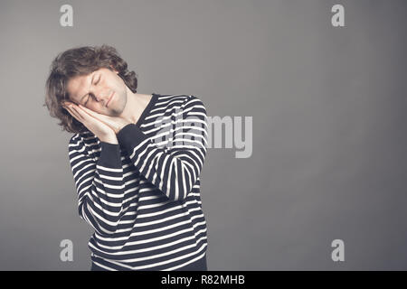 Giovane uomo in strisce in bianco e nero, maglione vuole dormire. Mettere la testa tra le mani e gli occhi chiusi mentre sorridente. In piedi su sfondo grigio. Foto Stock