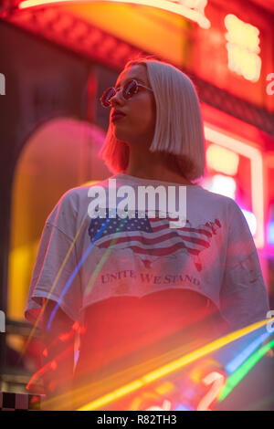 Ritratto cinematografico della ragazza bionda con il bianco di t-shirt e bandiera americana sul segno al neon di notte Foto Stock