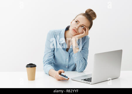 Ragazza dei sogni orologio si muovono più velocemente come sensazione annoiarsi in ufficio, testa pendente su palm ammirate angolo superiore sinistro indifferente seduta vicino laptop e tenendo lo smartphone, fantasticando, bere caffè Foto Stock