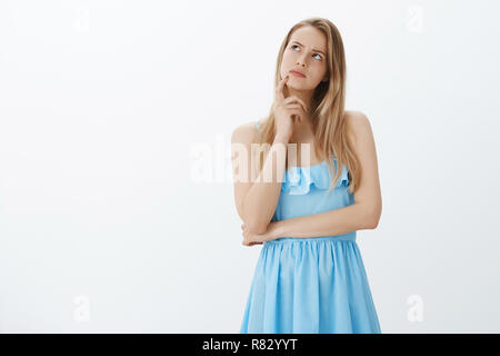 Ragazza ricordando la conversazione partendo overthink, sempre attivato. Riflessivo esitanti e focalizzato attraente giovane donna con capelli biondi in vestito blu alla ricerca nell'angolo superiore sinistro, pensando Foto Stock