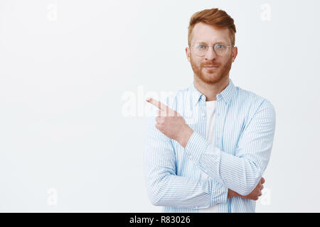 Guarda là ora. Ritratto di bello redhead maschio con la barba in eyewear e camicia rivolta ad angolo superiore sinistro e sorridente, stando in piedi in casual e prepotente pongono mentre a dare indicazioni ai dipendenti Foto Stock