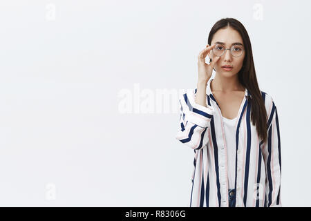 Inquadratura orizzontale di naturale attraente donna asiatica con pelle abbronzata e lunghi capelli scuri, toccando bicchieri arrotondati su gli occhi indossando elegante camicia a righe su t-shirt bianco, sguardo sognante in telecamera Foto Stock