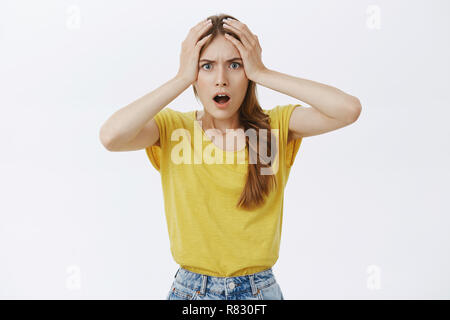 Ragazza avente un enorme disastro sul lavoro sentimento ansioso e orinato tenendo le mani sulla testa nel gesto disperato bocca di apertura dalla sorpresa accigliata standing insoddisfatto e tormentato oltre il muro grigio Foto Stock