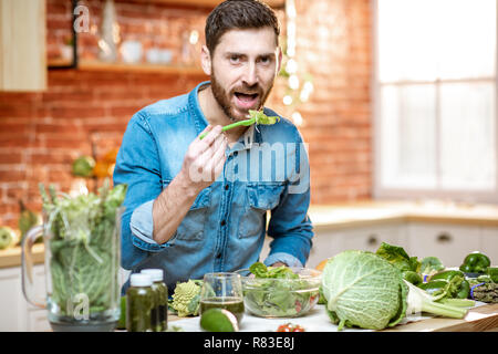 Uomo bello mangiare insalata sana seduta al tavolo pieno di ingredienti verde sulla cucina a casa Foto Stock