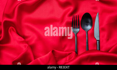 Il giorno di San Valentino la cena, tabella, posizionare l'impostazione. Posate, nero forchetta, cucchiaio e coltello su raso rosso sfondo, copia spazio, vista dall'alto. Foto Stock