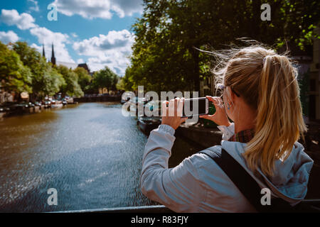 Vista posteriore del turista femminile prendendo la foto del canale di Amsterdam sul telefono cellulare sulla soleggiata giornata autunnale. Oro caldo pomeriggio di sole. Viaggi in Europa Foto Stock