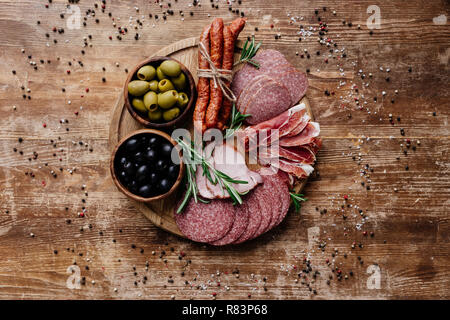 Vista superiore del round tagliere con deliziosi salami, il prosciutto e le olive in due ciotole sul tavolo di legno con spezie sparse Foto Stock