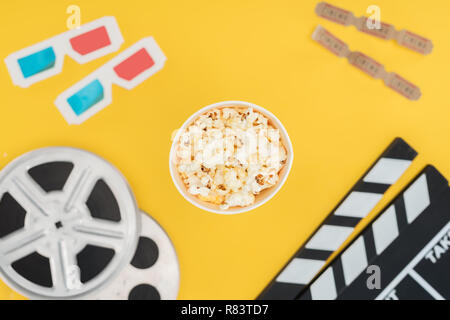 Vista superiore della clapperboard, bobine di pellicola, occhiali 3d, i biglietti del cinema e popcorn isolato della benna su giallo Foto Stock