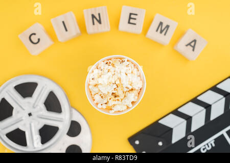 Vista superiore dei cubi di legno con 'cinema' scritte, clapperboard, bobine di pellicola e popcorn isolato della benna su giallo Foto Stock