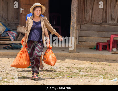 Thakhek, Laos - Aprile 20, 2018: donna locale che porta sacchetti pesanti nella parte anteriore di una casa in legno in un remoto villaggio rurale del sud Laos Foto Stock