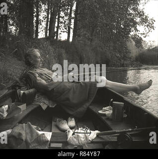 Degli anni Cinquanta, la signora anziana in scalza mentire indietro in una barca a remi in corrispondenza di un bordo del fiume in possesso di una canna da pesca, Inghilterra, Regno Unito. Foto Stock