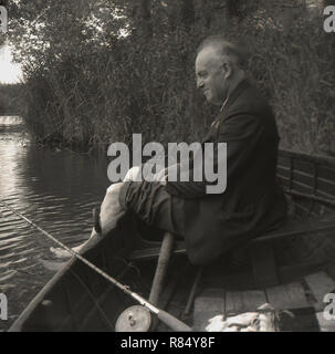 Degli anni Cinquanta, un uomo di mezza età che indossa un abito seduto su una barca a remi da un bordo del fiume con i suoi pantaloni laminati per le sue ginocchia Dondolando i suoi piedi in acqua, Inghilterra, Regno Unito. La sua canna da pesca è accanto a lui sulla barca. Foto Stock