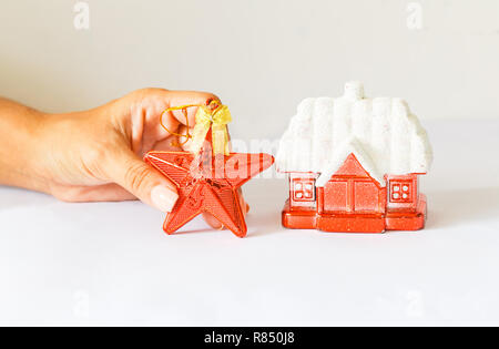 Anno nuovo decorazione rossa rifugio con una coperta di neve tetto e una stella rossa su uno sfondo bianco. femmina di mano che tiene una stella. Foto Stock