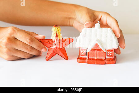 Anno nuovo decorazione rossa rifugio con una coperta di neve tetto e una stella rossa su uno sfondo bianco. femmina di mano che tiene una stella. Foto Stock