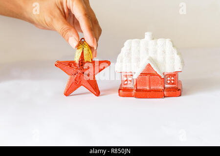Anno nuovo decorazione rossa rifugio con una coperta di neve tetto e una stella rossa su uno sfondo bianco. femmina di mano che tiene una stella. Foto Stock