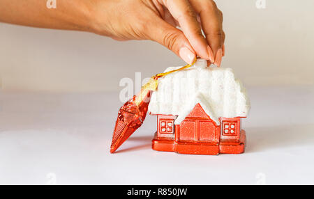 Anno nuovo decorazione rossa rifugio con una coperta di neve tetto e una stella rossa su uno sfondo bianco. femmina di mano che tiene una stella. Foto Stock