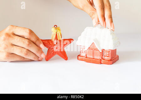 Anno nuovo decorazione rossa rifugio con una coperta di neve tetto e una stella rossa su uno sfondo bianco. femmina di mano che tiene una stella. Foto Stock