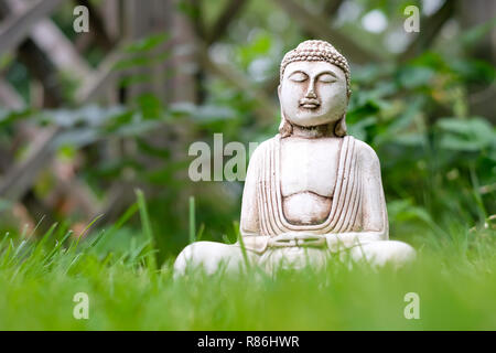 Di piccole dimensioni e di colore bianco statua del Buddha in una meditazione posano con erba verde in primo piano e il brillante naturale sfondo sfocato. Simbolo religioso del Buddismo Foto Stock